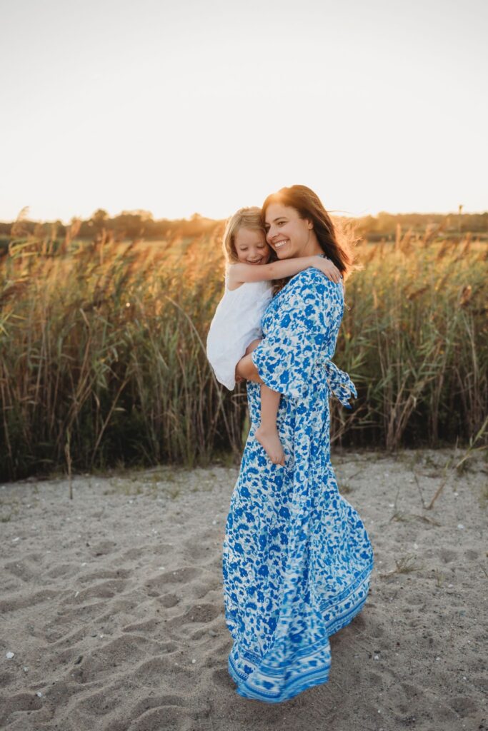 A mother in a flowing blue floral dress holding her young daughter during a golden hour outdoor session in a grassy field at sunset