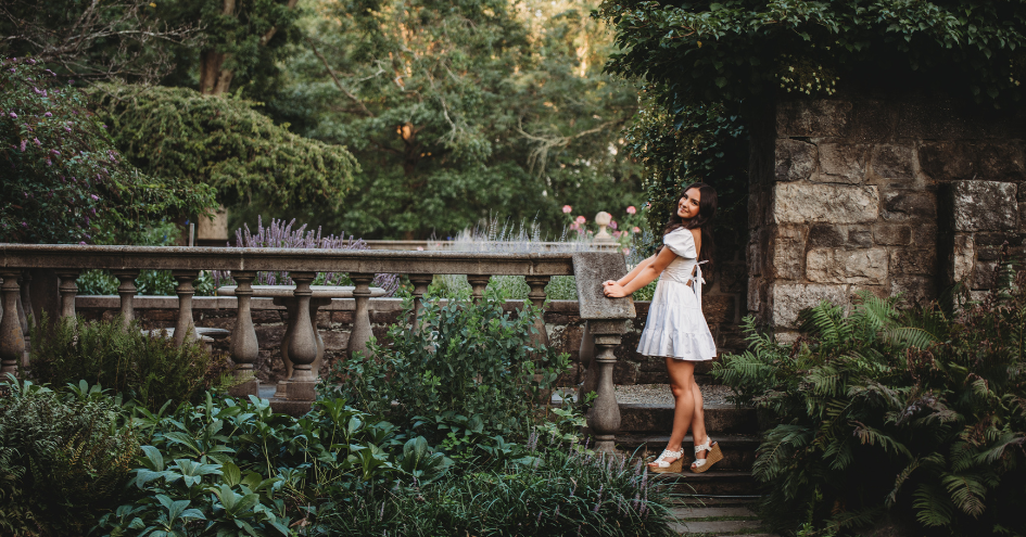 High school senior girl in white dress posing on stone steps in a lush garden in Connecticut