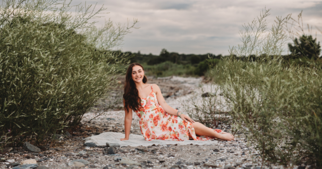 Senior girl in a white and orange floral dress sitting on a beach blanket surrounded by greenery