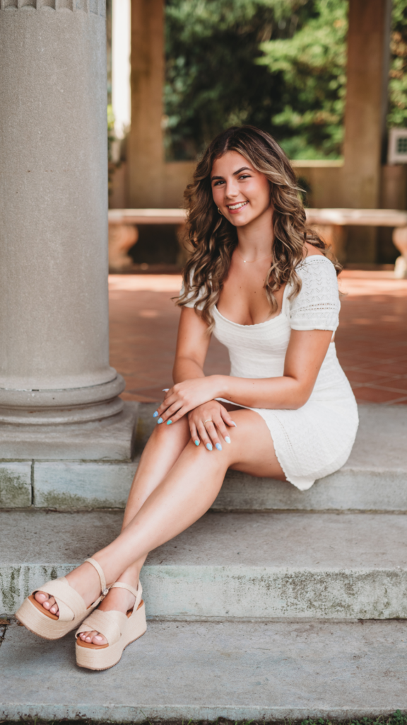High school senior girl in white dress posing on stone steps in a lush garden in Connecticut