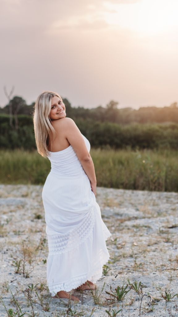 High school senior girl in a white sundress smiling over her shoulder during a sunset beach session