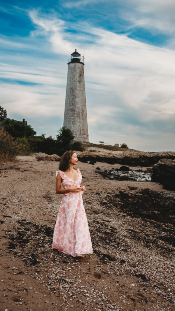 High school senior girl in pink an white dress walking near a lighthouse in Connecticut
